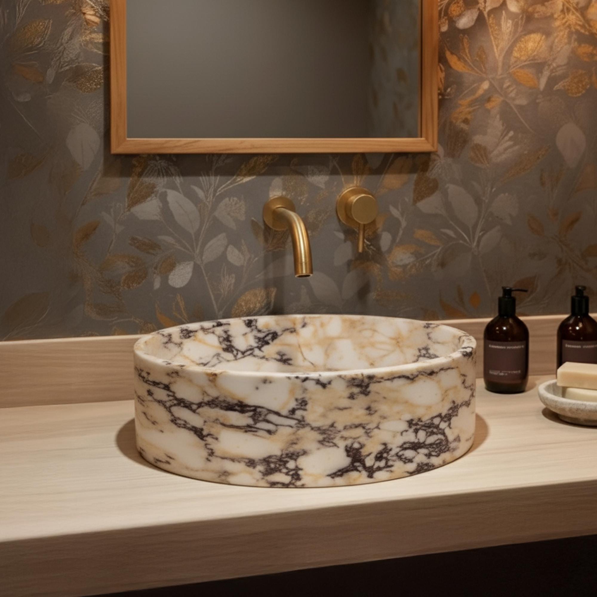 A round Calacatta Amber Viola marble vessel sink sits on a light wood vanity, paired with a brushed gold wall-mounted faucet.