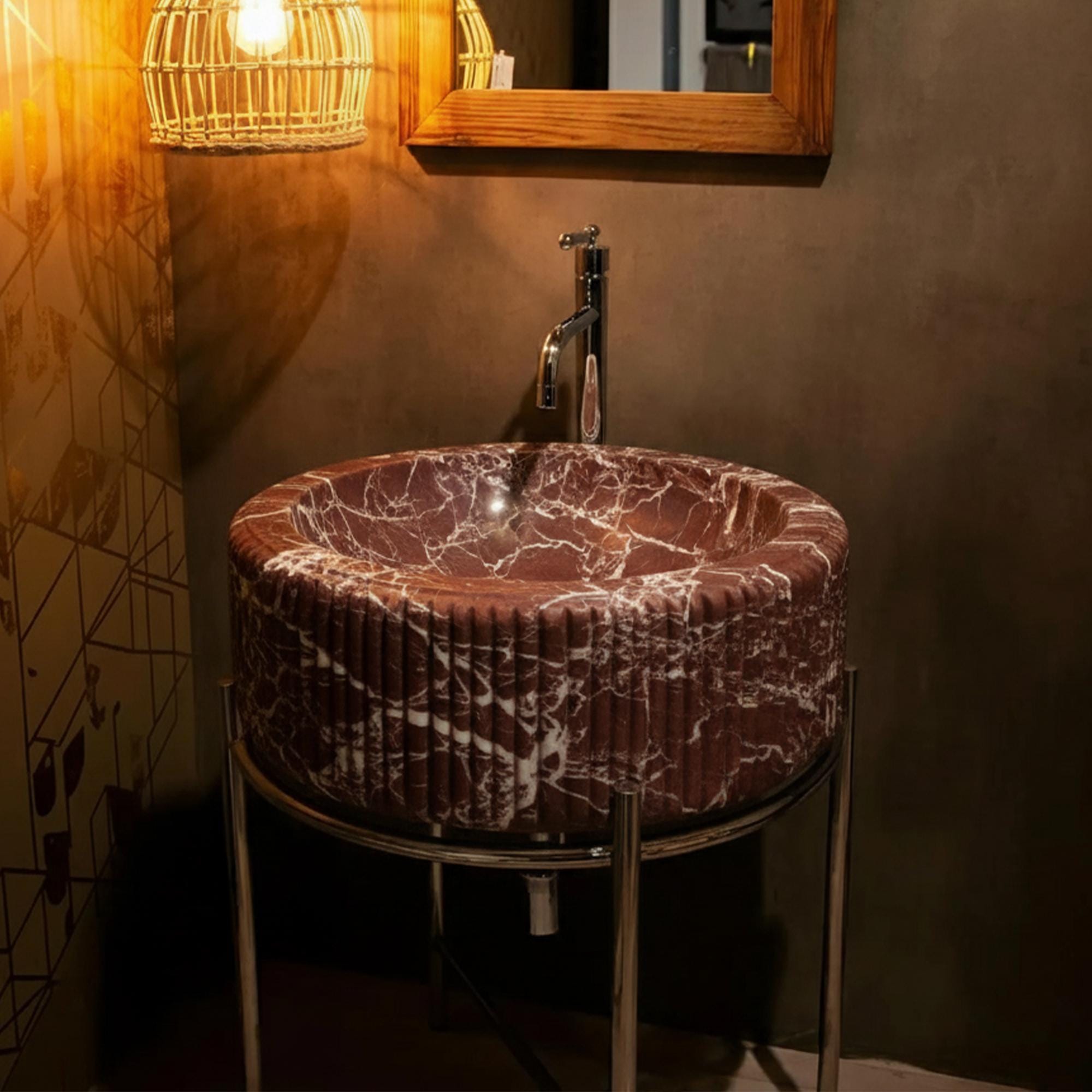 This image shows a fluted round sink made from Rosso Levanto marble, paired with a sleek chrome base and moody lighting.
