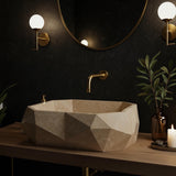 A bold geometric travertine vessel sink sits on a wood vanity with gold fixtures and soft candlelight ambiance.